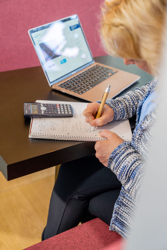 Student is zittend haar rekentoets pabo aan het voorbereiden. Ze heeft een blauw vest aan. De hulpmiddelen die ze gebruikt zijn een rekenmachine, een kladblok en haar laptop